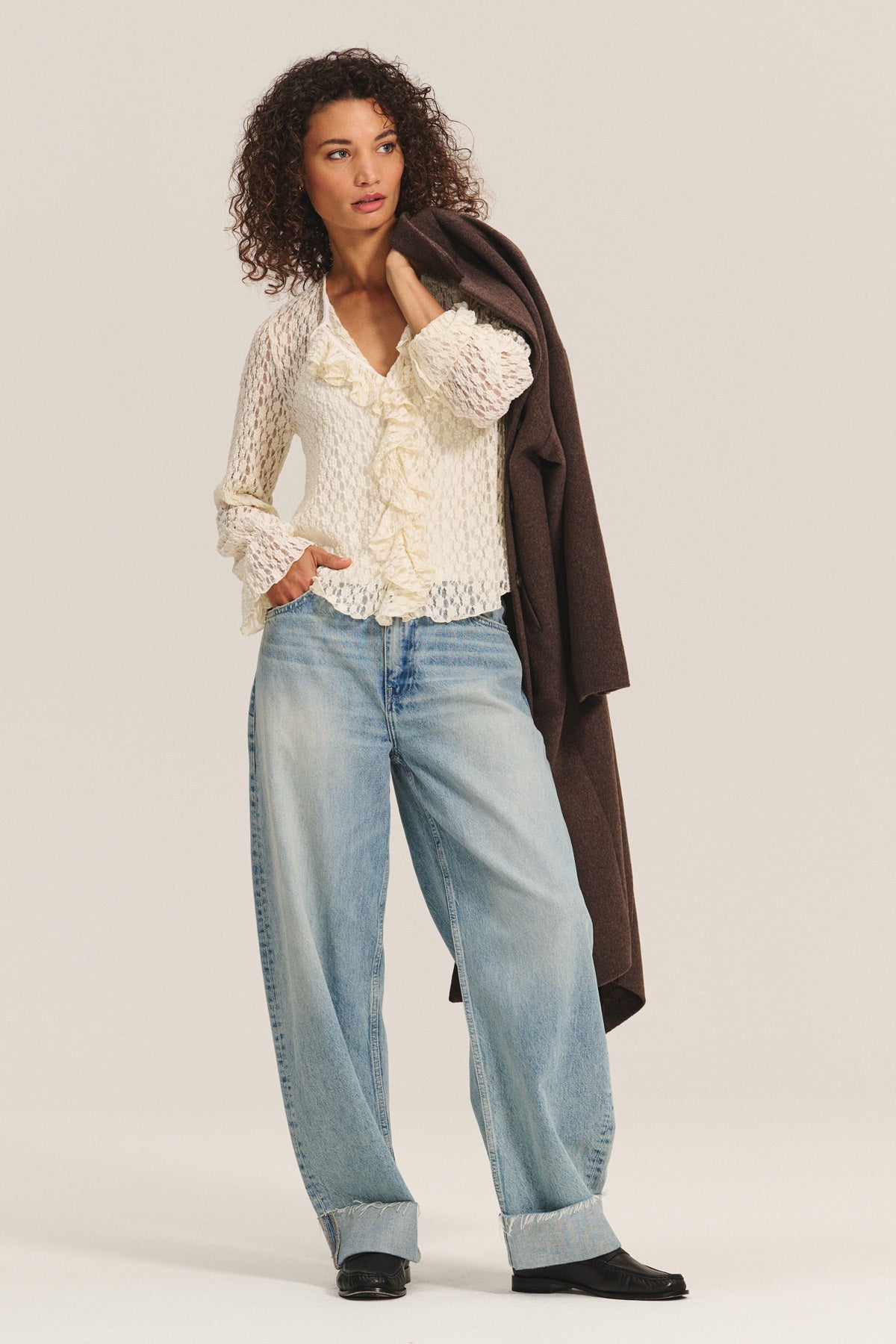 A woman stands with a brown coat over her shoulder, wearing the NATALIE TOP by Velvet by Graham & Spencer—a cream ruffled blouse—paired with loose, cuffed blue jeans and black shoes.-40263657291969