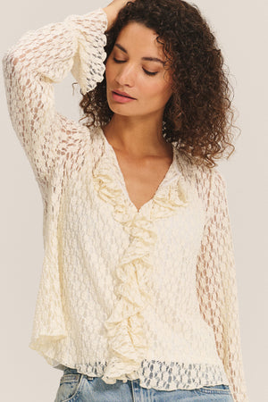 A woman poses gracefully with one arm raised and eyes closed, wearing the NATALIE TOP by Velvet by Graham & Spencer—a cream-colored cotton lace top with ruffle details and long sleeves—paired with light blue jeans.