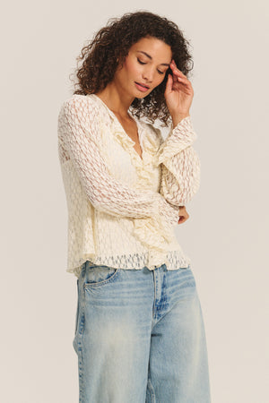 A woman wears the NATALIE TOP by Velvet by Graham & Spencer, a cream-colored cotton lace blouse with delicate ruffles, paired with light blue jeans, standing against a plain backdrop.