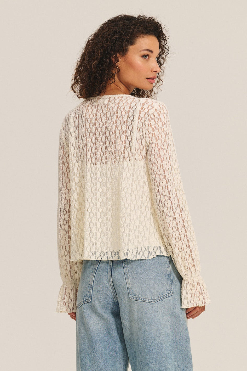 A woman stands with her back to the camera, wearing the NATALIE TOP by Velvet by Graham & Spencer—a sheer, long-sleeve cotton lace top with ruffle details—paired with light blue jeans against a plain background.