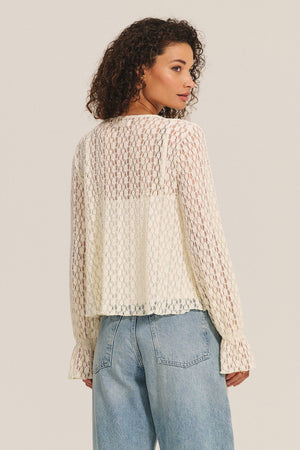 A woman stands with her back to the camera, wearing the NATALIE TOP by Velvet by Graham & Spencer—a sheer, long-sleeve cotton lace top with ruffle details—paired with light blue jeans against a plain background.