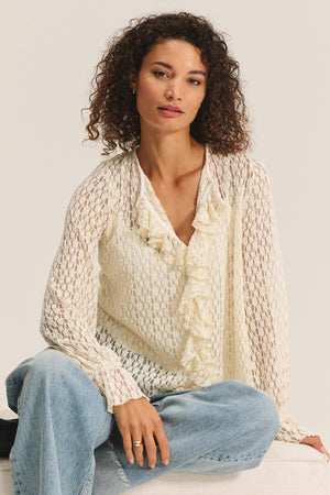 A woman with curly hair wears the NATALIE TOP by Velvet by Graham & Spencer—a cotton lace top with ruffle details—paired with light blue jeans, sitting with one knee raised against a plain background.