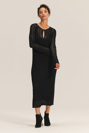 A woman wears the TABITHA DRESS by Velvet by Graham & Spencer, a black long-sleeve knit midi-to-maxi dress with a keyhole neckline, paired with black ankle boots, standing against a plain light background.