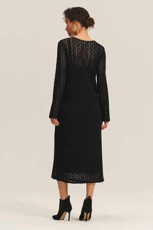 A woman faces away, wearing the Velvet by Graham & Spencer TABITHA DRESS— a long-sleeve, stretch cotton lace dress in midi-to-maxi length—styled with black heeled ankle boots against a plain background.