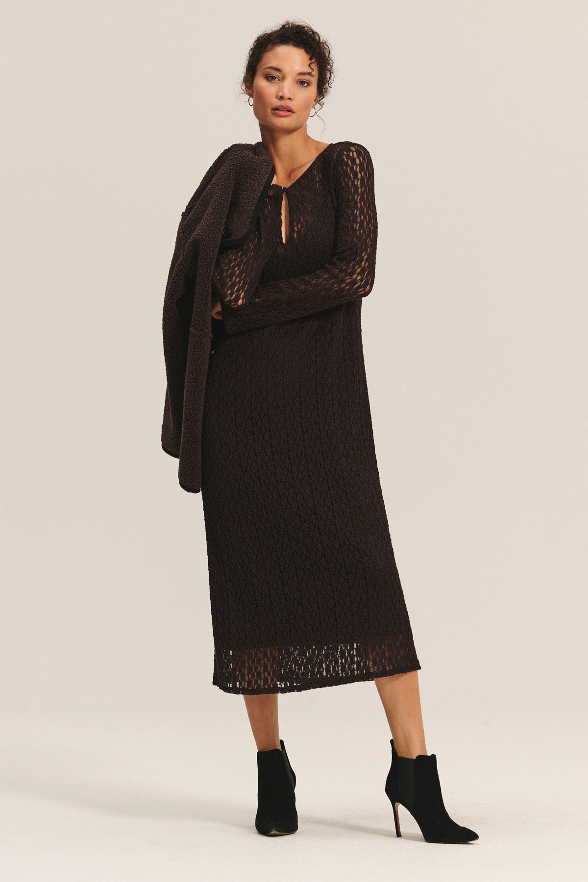 A woman stands against a plain background wearing the TABITHA DRESS by Velvet by Graham & Spencer—an ankle-length, long-sleeve brown dress—paired with black ankle boots and holding a matching brown garment over her shoulder.-40263670169793