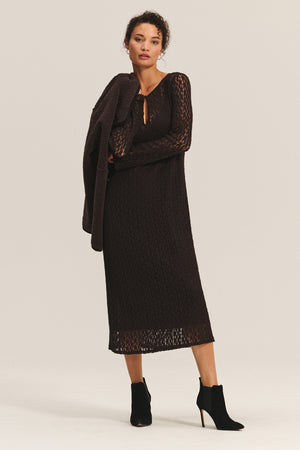A woman stands against a plain background wearing the TABITHA DRESS by Velvet by Graham & Spencer—an ankle-length, long-sleeve brown dress—paired with black ankle boots and holding a matching brown garment over her shoulder.