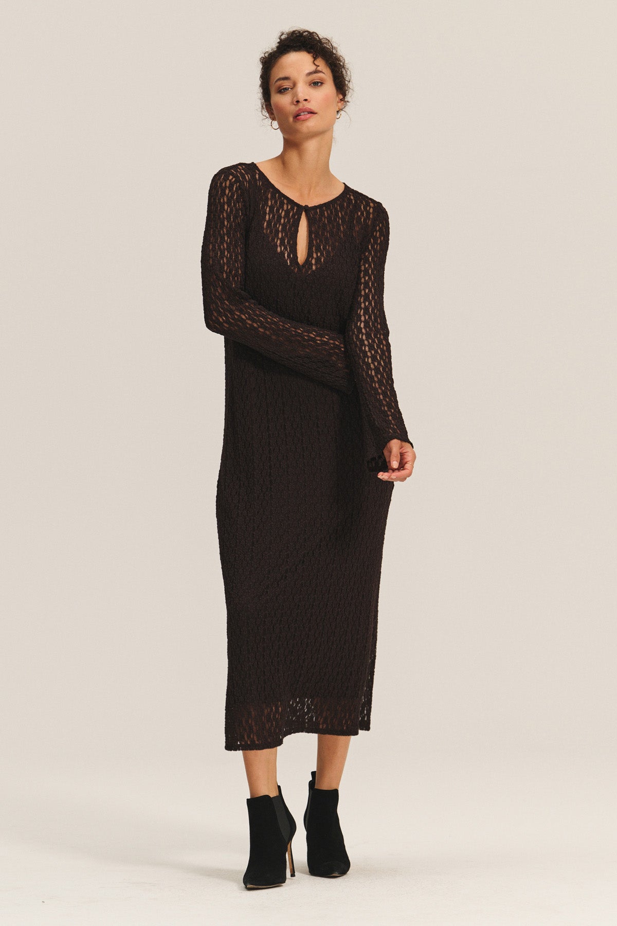 A woman wears the TABITHA DRESS by Velvet by Graham & Spencer—a long-sleeve, black, semi-sheer knit midi-to-maxi dress with a keyhole neckline—paired with black ankle boots against a plain light background.-40263670137025