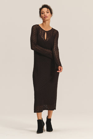 A woman wears the TABITHA DRESS by Velvet by Graham & Spencer—a long-sleeve, black, semi-sheer knit midi-to-maxi dress with a keyhole neckline—paired with black ankle boots against a plain light background.