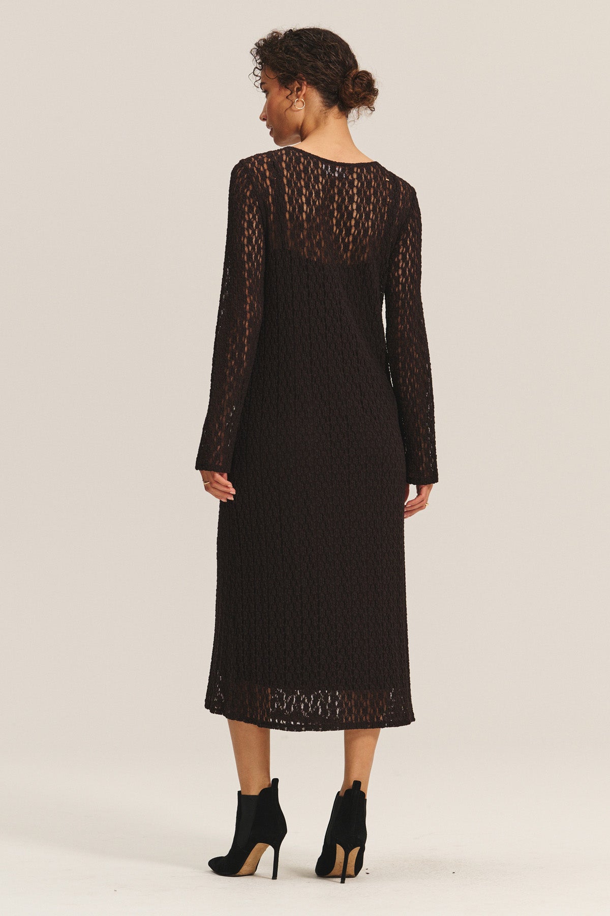 A woman faces away, wearing the TABITHA DRESS by Velvet by Graham & Spencer—a black, long-sleeve cotton lace midi dress—paired with black heeled ankle boots against a plain background.-40263670104257