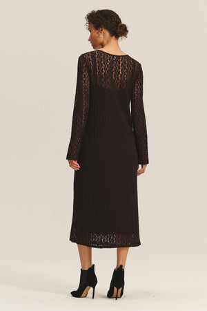 A woman faces away, wearing the TABITHA DRESS by Velvet by Graham & Spencer—a black, long-sleeve cotton lace midi dress—paired with black heeled ankle boots against a plain background.