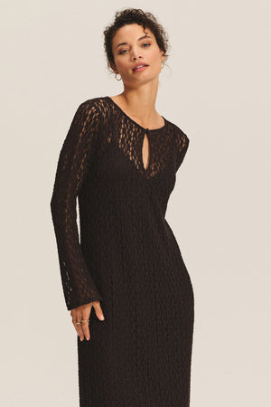 Someone wearing the TABITHA DRESS by Velvet by Graham & Spencer—a long-sleeve, black textured midi-to-maxi dress with a small keyhole cutout at the neckline—stands against a plain background.