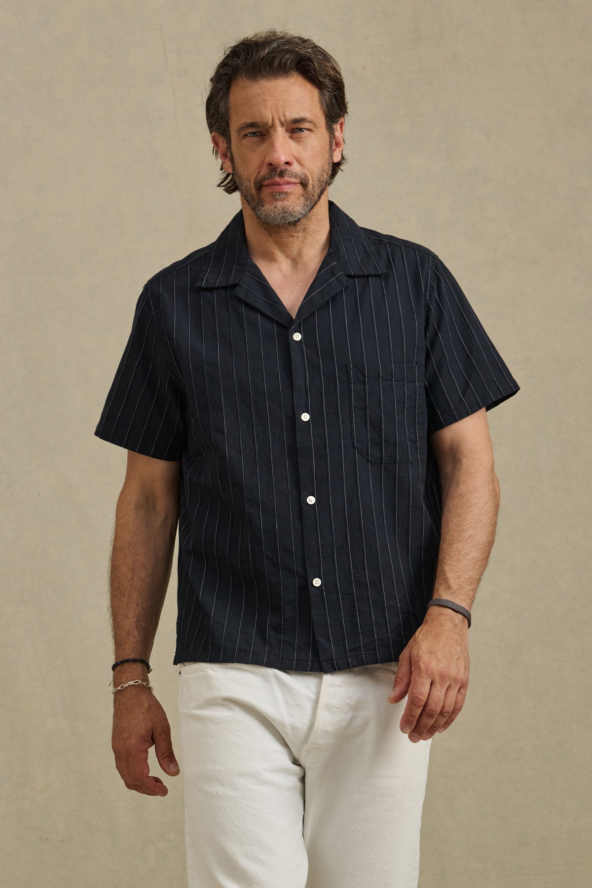 A man with short brown hair and a beard wears the black GUNNER SHIRT by Velvet by Graham & Spencer, featuring subtle pinstripes, paired with white pants against a plain beige background.