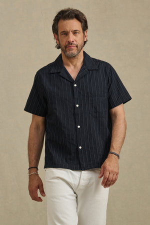 A man with short brown hair and a beard wears the black GUNNER SHIRT by Velvet by Graham & Spencer, featuring subtle pinstripes, paired with white pants against a plain beige background.