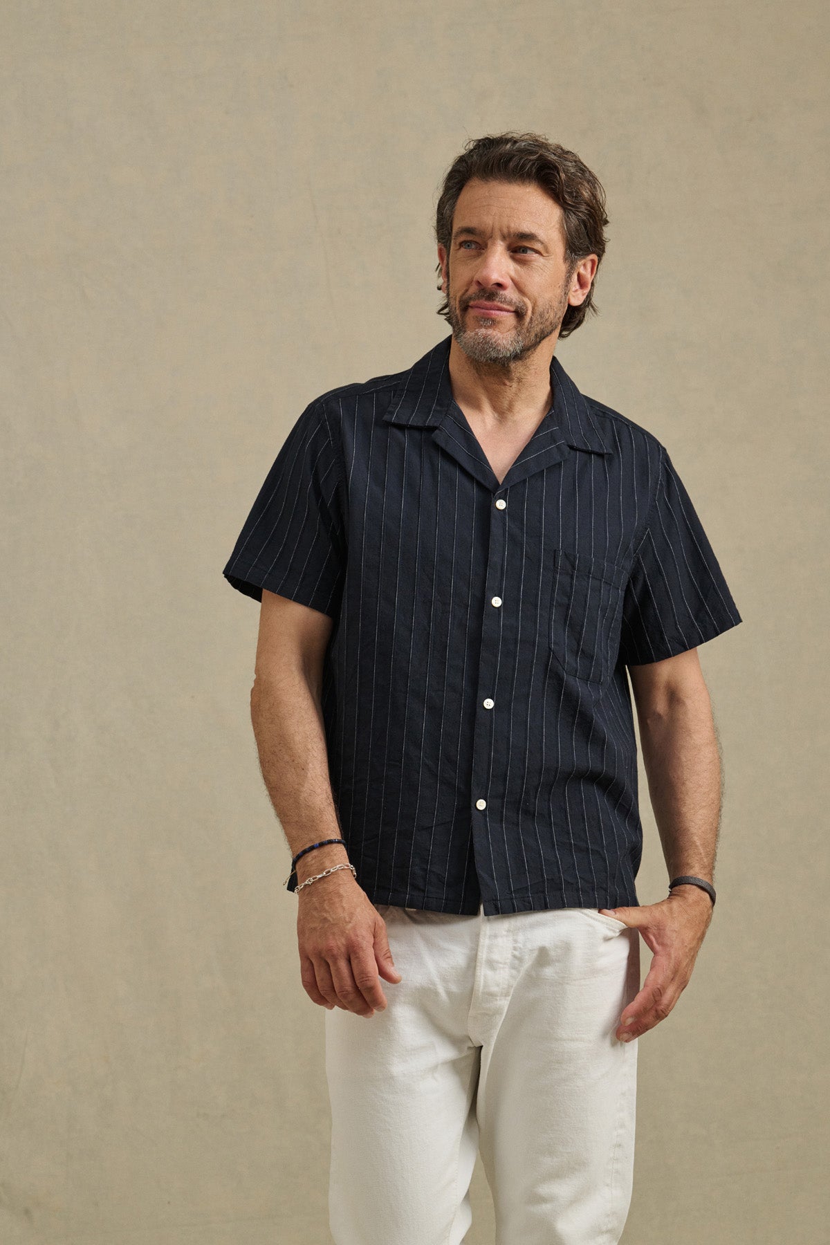 A man wears the GUNNER SHIRT by Velvet by Graham & Spencer in dark stripe dobby cotton with white pants, standing against a plain beige background.