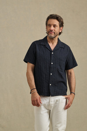 A man wears the GUNNER SHIRT by Velvet by Graham & Spencer in dark stripe dobby cotton with white pants, standing against a plain beige background.