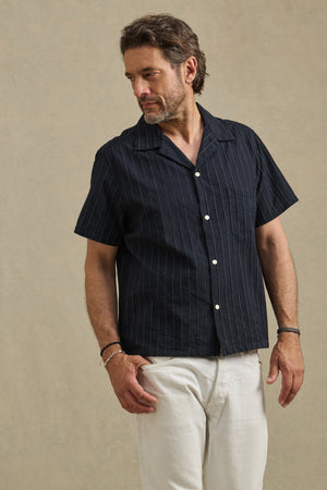 A man stands against a plain beige background, looking to his left, wearing the Velvet by Graham & Spencer GUNNER SHIRT—short-sleeved with a camp collar in stripe dobby cotton—and white pants.