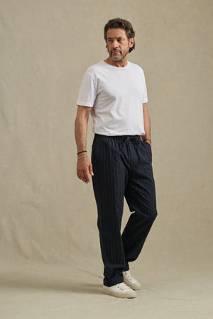 A man stands against a plain background, wearing Velvet by Graham & Spencer's SAINT PANT, a white t-shirt, and white sneakers, with one hand in his pocket.
