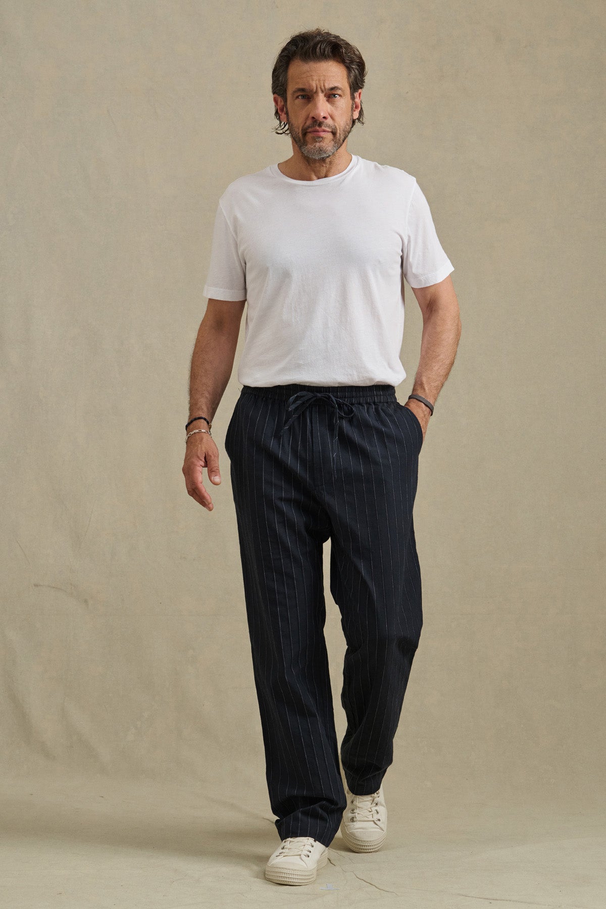 A man stands against a plain background, wearing Velvet by Graham & Spencer's SAINT PANT in dark pinstripes, paired with a white t-shirt and white sneakers.