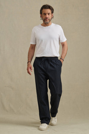 A man stands against a plain background, wearing Velvet by Graham & Spencer's SAINT PANT in dark pinstripes, paired with a white t-shirt and white sneakers.