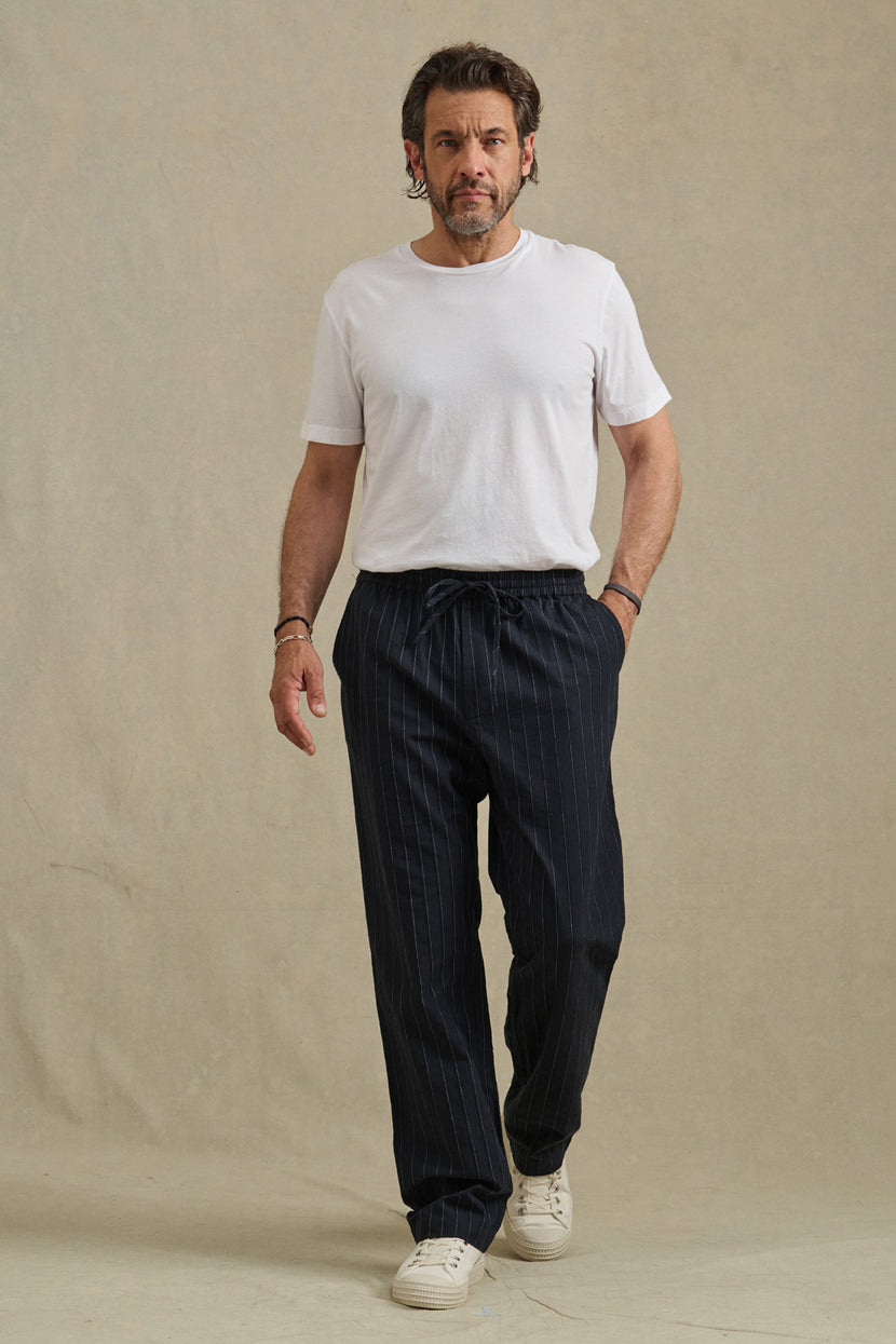 A man stands against a plain background, wearing Velvet by Graham & Spencer's SAINT PANT in dark pinstripes, paired with a white t-shirt and white sneakers.