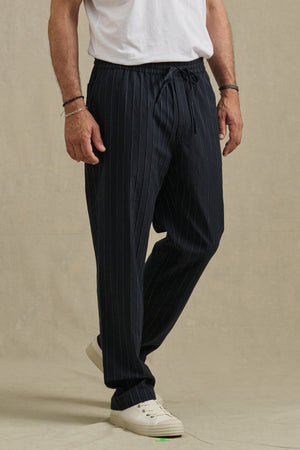 A person wearing Velvet by Graham & Spencer's SAINT PANT stands against a plain beige background, paired with a white t-shirt and white sneakers.