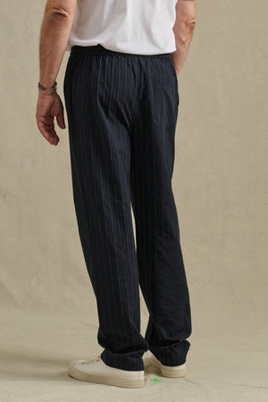 A person is seen from the back standing on a light floor, wearing a white T-shirt, Velvet by Graham & Spencer SAINT PANT in black pinstripe, and white sneakers.