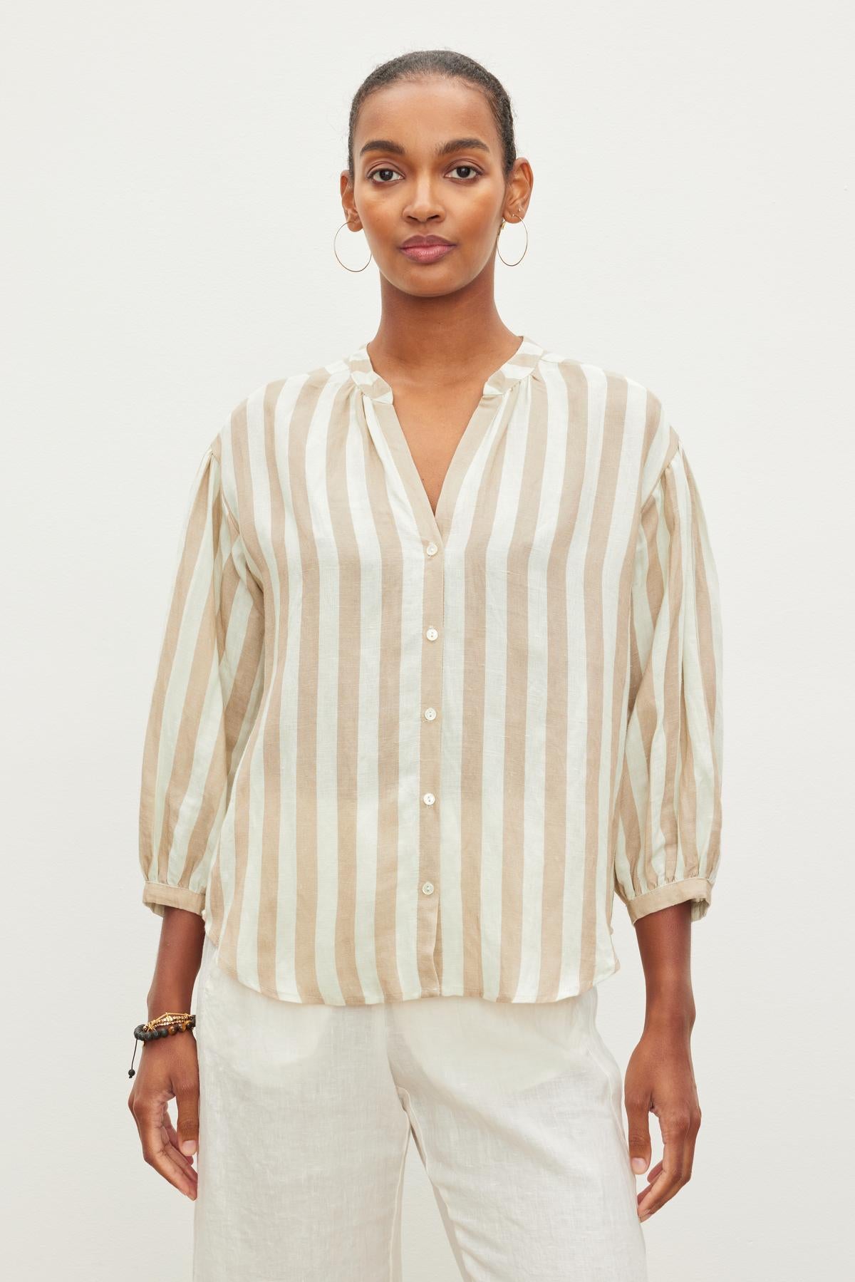   A person stands against a plain white background, wearing a GABBY STRIPED LINEN TOP by Velvet by Graham & Spencer with rolled-up sleeves and white pants. 