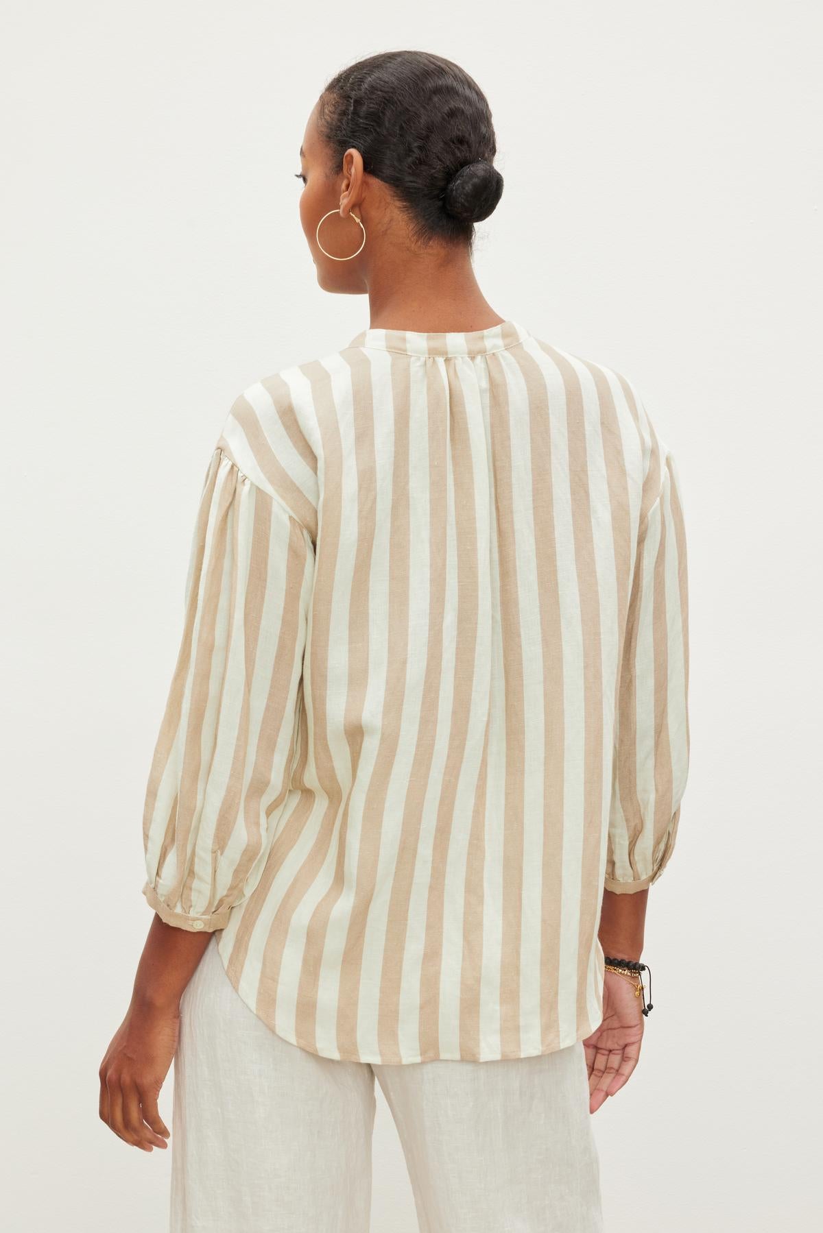   A person with a neat bun hairstyle wearing a GABBY STRIPED LINEN TOP by Velvet by Graham & Spencer and white pants is standing with their back facing the camera. 