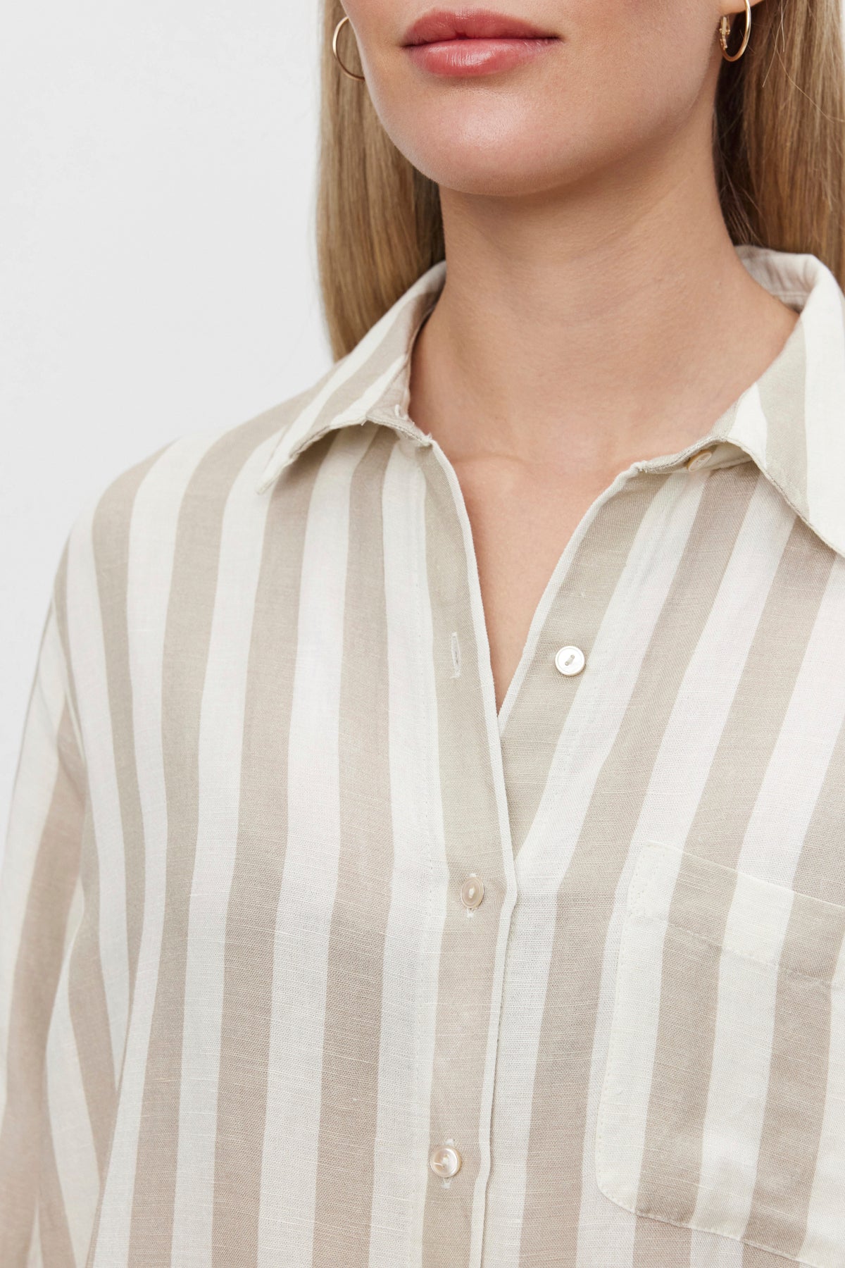   A person wearing the HARLOW STRIPED LINEN BUTTON-UP SHIRT by Velvet by Graham & Spencer. The beige and white vertically striped shirt features a collar, and its relaxed fit complements the timeless stripes perfectly. The person's face is partially visible, with focus on the upper torso and shirt details. 