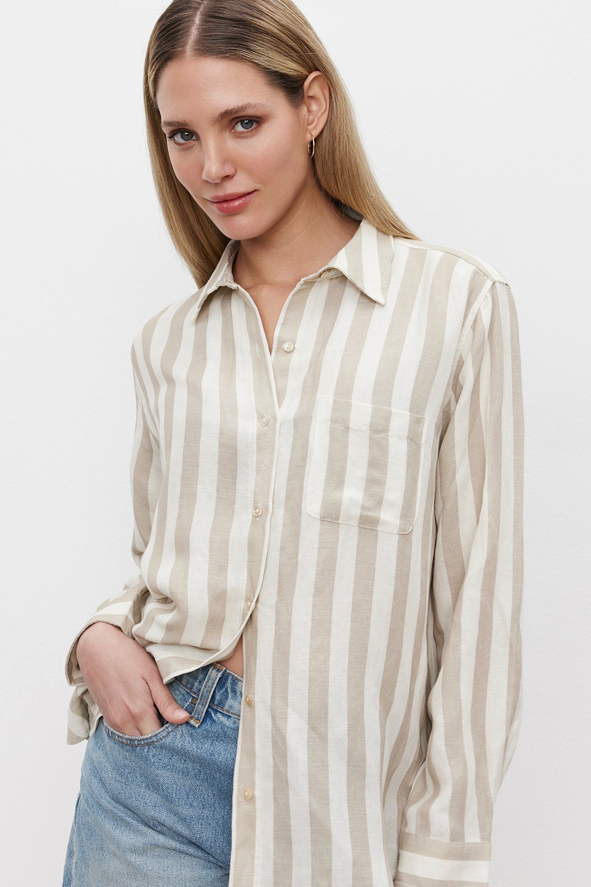   A woman with long blonde hair wearing a relaxed fit, beige and white HARLOW STRIPED LINEN BUTTON-UP SHIRT from Velvet by Graham & Spencer, paired with blue jeans, stands against a plain white background. 