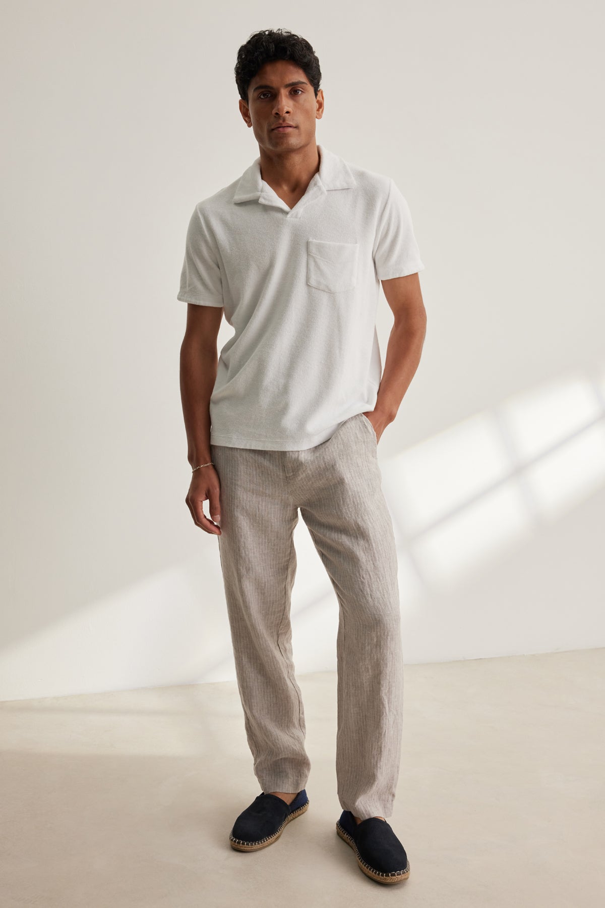 A man stands indoors against a plain light background wearing a white short-sleeve polo, Velvet by Graham & Spencer's JAE LINEN PANT with stripes and an elastic waistband, and dark slip-on shoes.-39152698228929