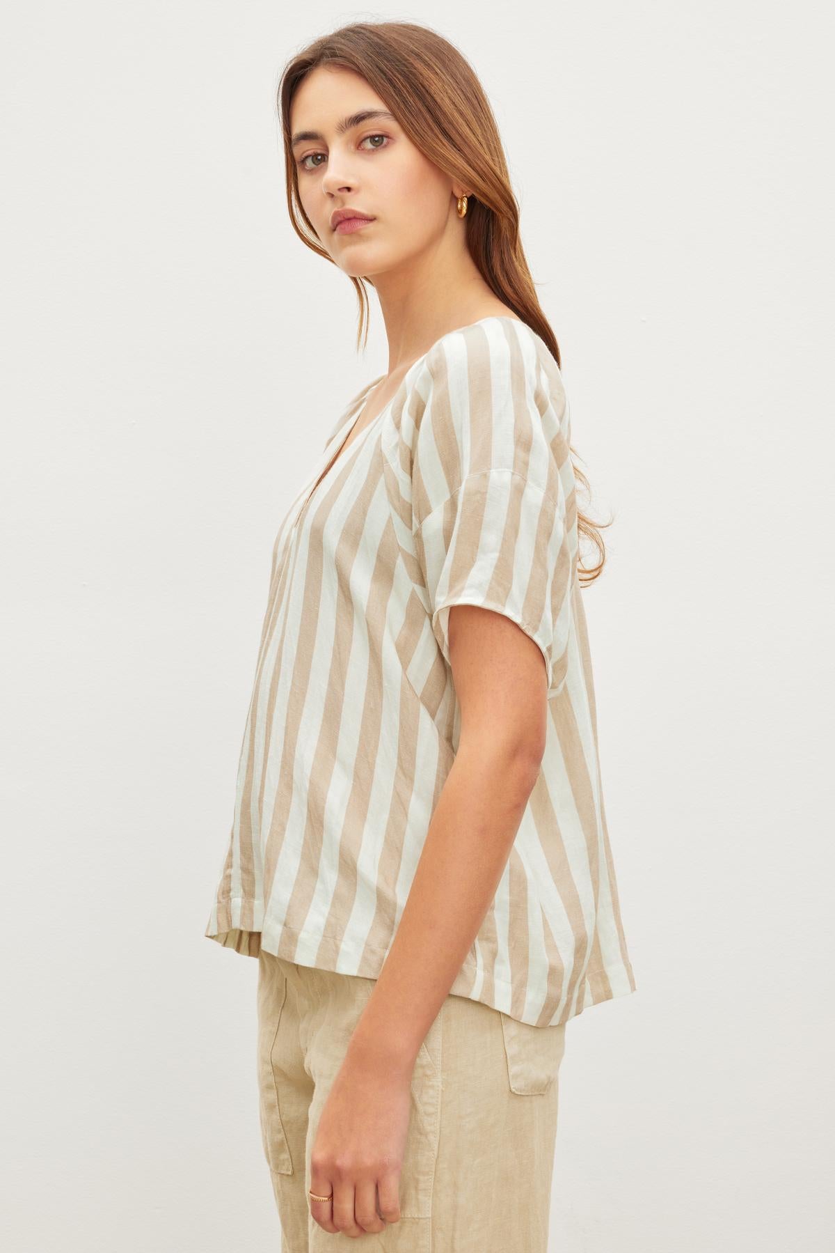   A person with long hair stands against a plain background, wearing the Velvet by Graham & Spencer KATY STRIPED LINEN TOP and relaxed fit beige pants, evoking effortless West Coast vibes. 