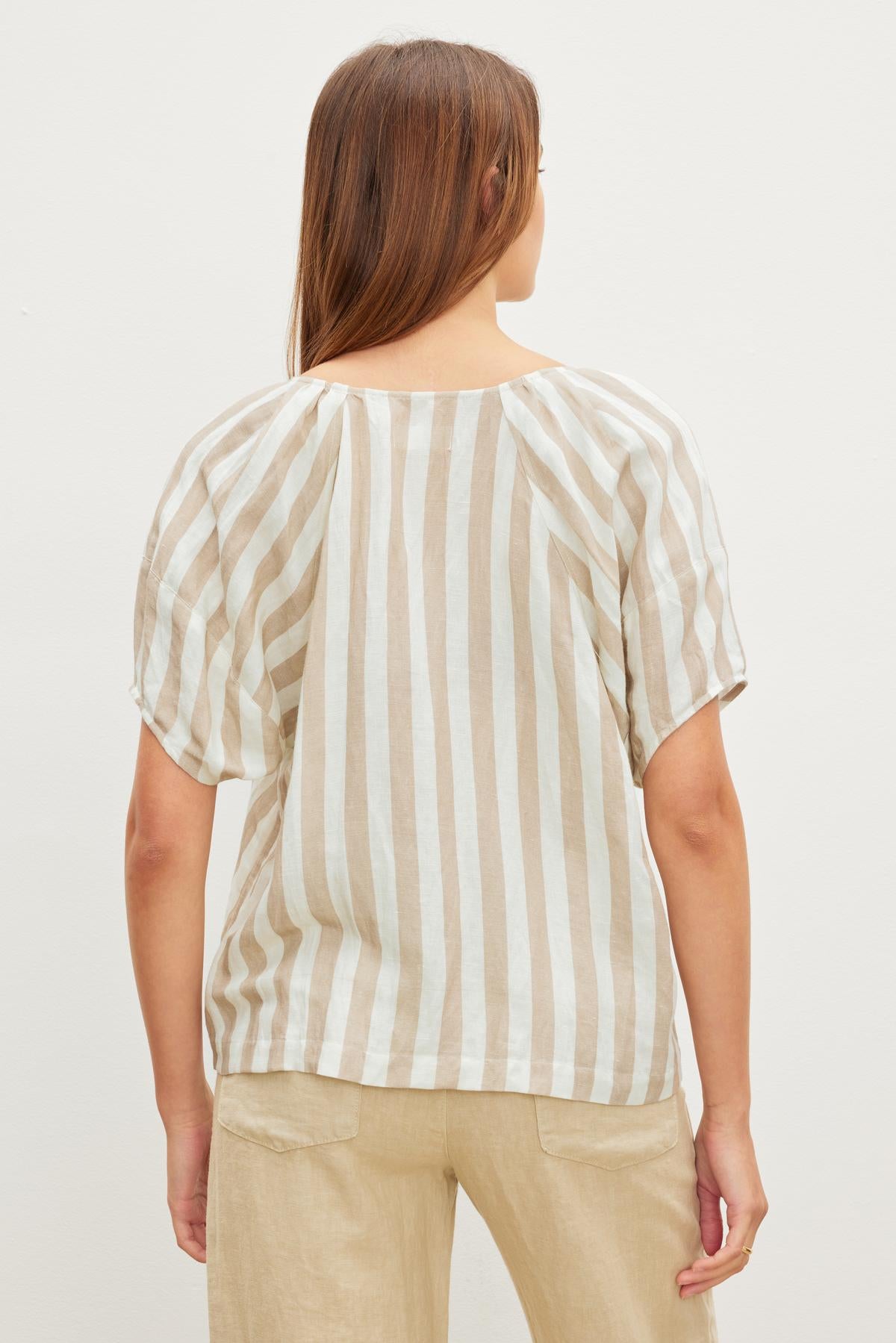   A person with long brown hair is standing with their back to the camera, wearing a beige and white striped short-sleeve Velvet by Graham & Spencer KATY STRIPED LINEN TOP with a v-neckline and relaxed fit beige pants. 