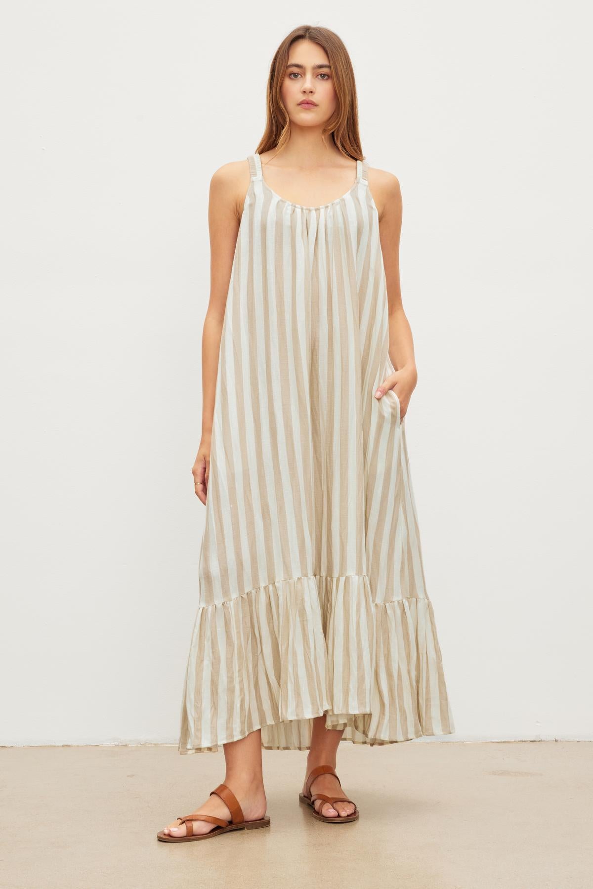   A person stands against a plain backdrop, wearing the MERADITH STRIPED LINEN MAXI DRESS by Velvet by Graham & Spencer. The sleeveless maxi dress features vertical beige and white stripes along with a ruffled hem, paired with brown sandals. Made of a woven linen blend, the dress adds an elegant yet laid-back vibe. 