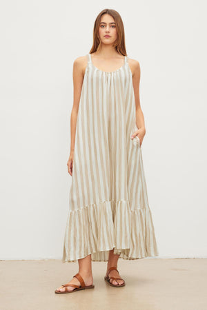 A person stands against a plain backdrop, wearing the MERADITH STRIPED LINEN MAXI DRESS by Velvet by Graham & Spencer. The sleeveless maxi dress features vertical beige and white stripes along with a ruffled hem, paired with brown sandals. Made of a woven linen blend, the dress adds an elegant yet laid-back vibe.