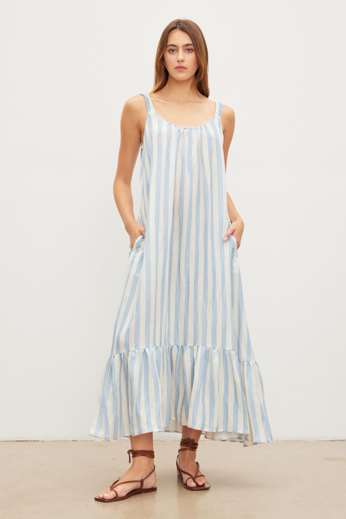   A woman stands against a plain background wearing a Velvet by Graham & Spencer MERADITH STRIPED LINEN MAXI DRESS with ruffle hem and brown sandals. 