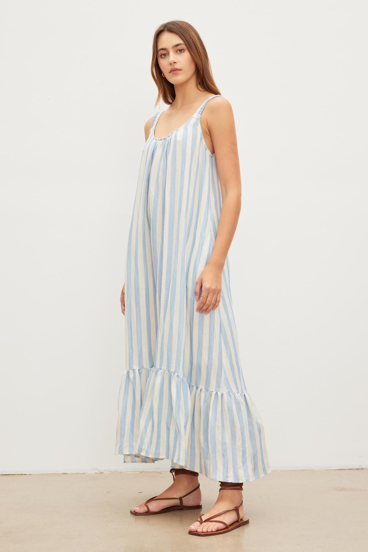   A woman stands against a plain background, wearing a Velvet by Graham & Spencer MERADITH STRIPED LINEN MAXI DRESS with thin straps and ruffled hemline, paired with brown sandals. 