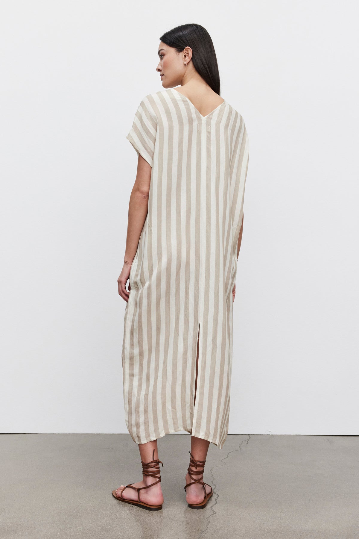 Woman standing in a profile view wearing a long beige and white striped Milla Striped Linen Kaftan Dress by Velvet by Graham & Spencer with a split hem, paired with brown sandals.-36247892656321