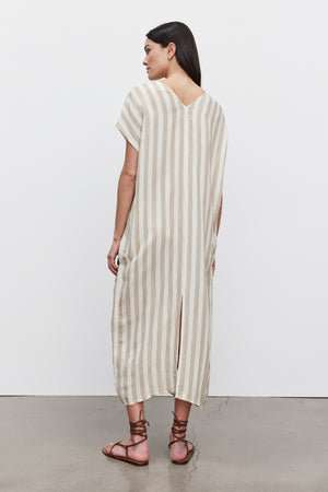 Woman standing in a profile view wearing a long beige and white striped Milla Striped Linen Kaftan Dress by Velvet by Graham & Spencer with a split hem, paired with brown sandals.