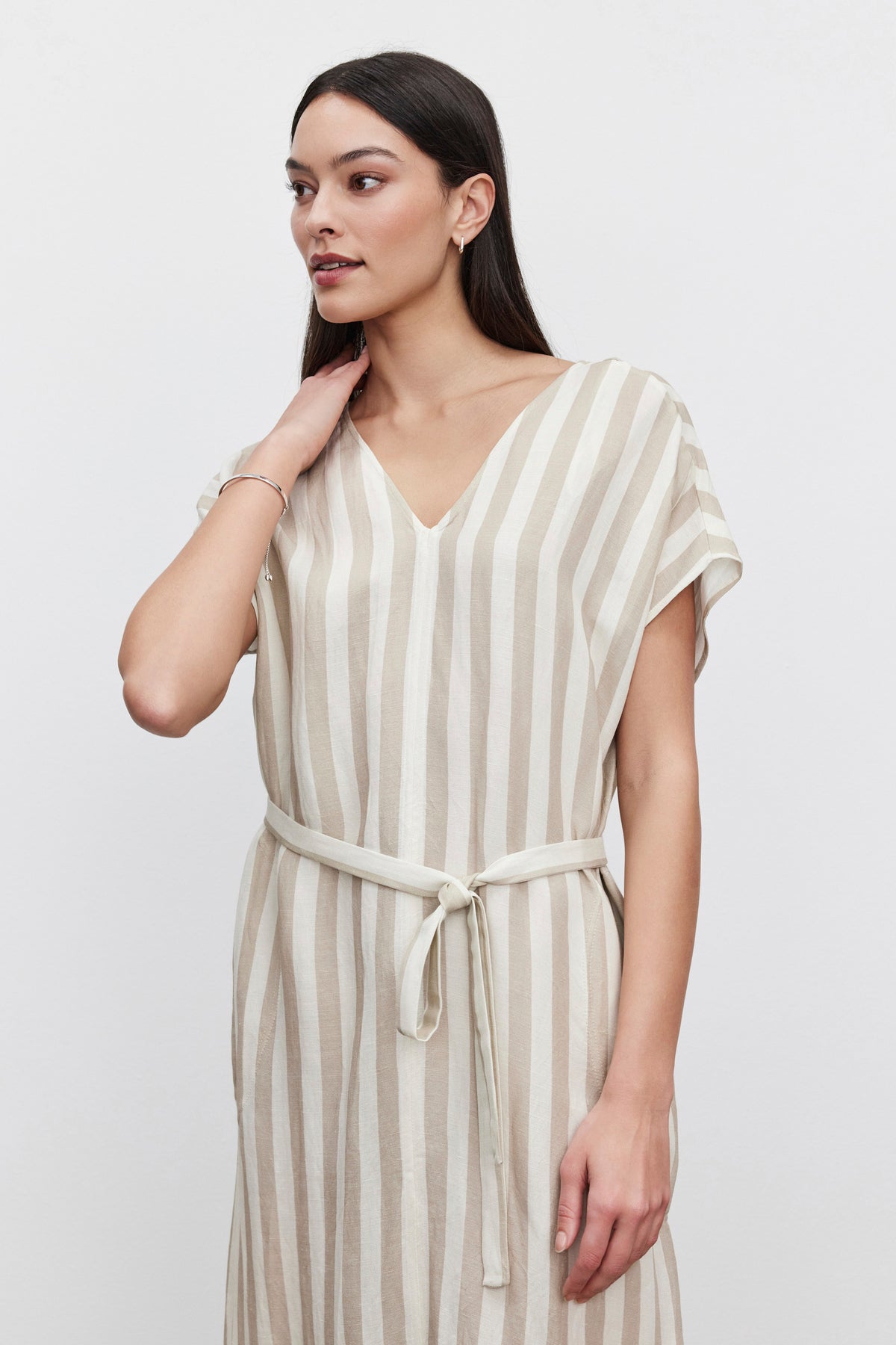   A woman in a MILLA STRIPED LINEN KAFTAN DRESS by Velvet by Graham & Spencer, with a tie waist, standing against a white background, touches her neck and looks to the side. 