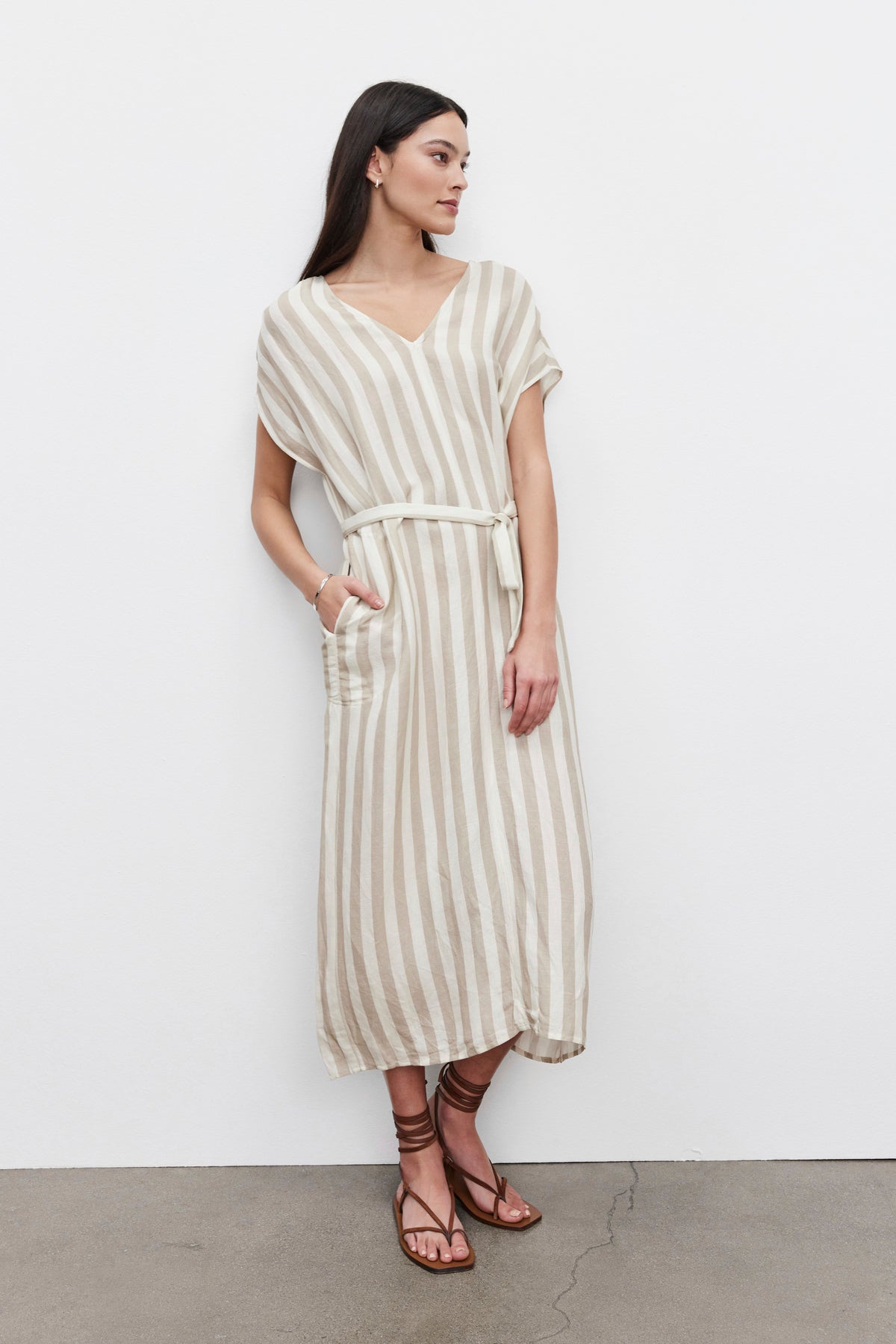   A woman stands against a white wall, wearing a Velvet by Graham & Spencer MILLA STRIPED LINEN KAFTAN DRESS with a belt and brown sandals. 