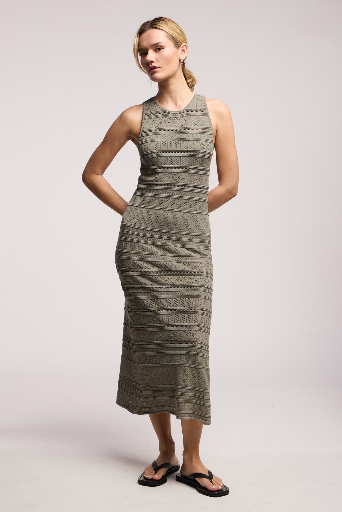 A woman wears the YLENA DRESS by Velvet by Graham & Spencer, a fitted olive sleeveless midi dress with textured horizontal patterns and an A-line silhouette, styled with black flip-flops against a plain background.