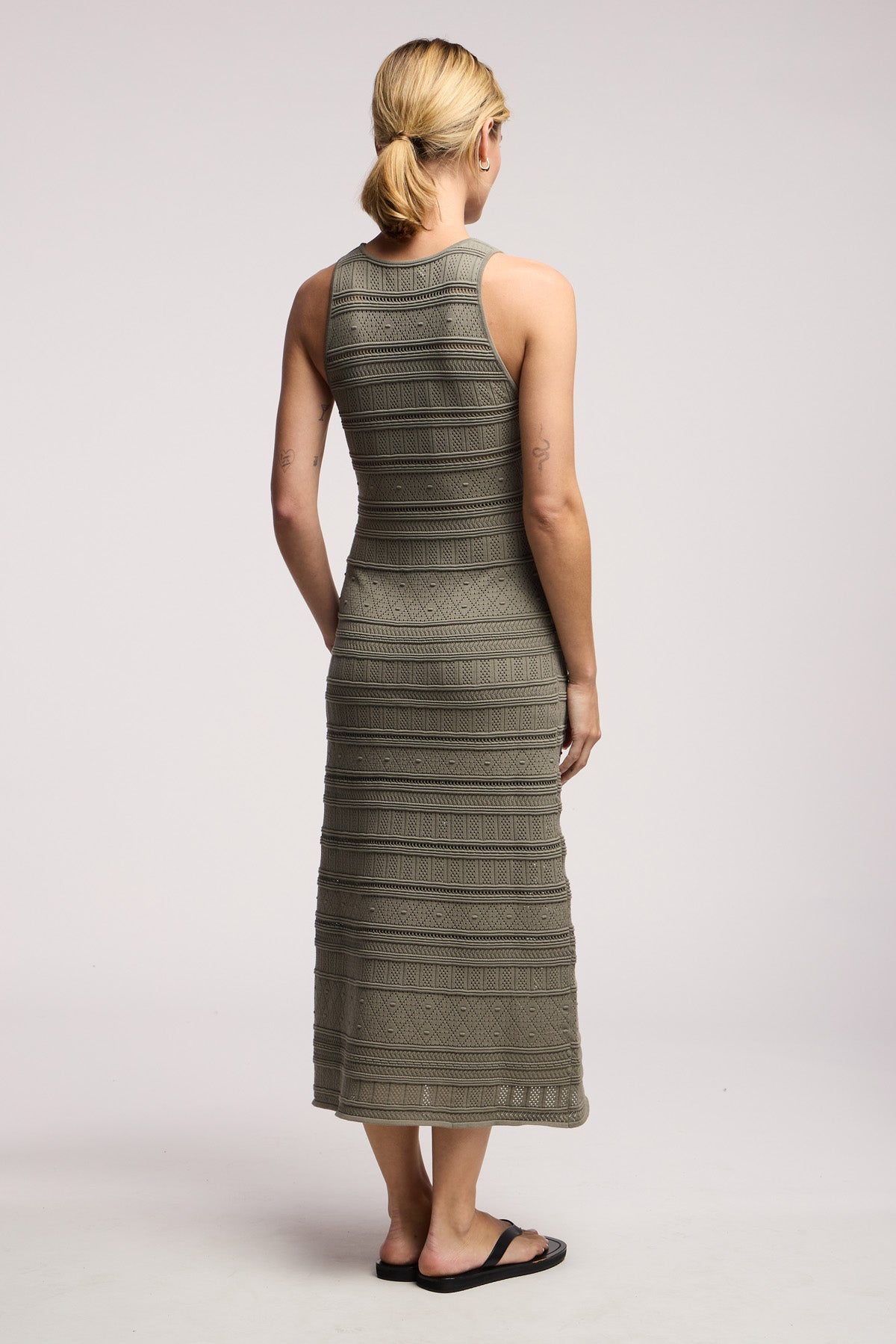 A barefoot person with blonde hair in a low ponytail stands facing away, wearing the YLENA DRESS by Velvet by Graham & Spencer—an olive green, sleeveless stripe pointelle midi dress with horizontal textured patterns.-41645211812033