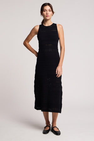 A woman stands against a plain background wearing the YLENA DRESS by Velvet by Graham & Spencer, a sleeveless textured midi with a fitted A-line silhouette, paired with black Mary Jane shoes for an elegant look.