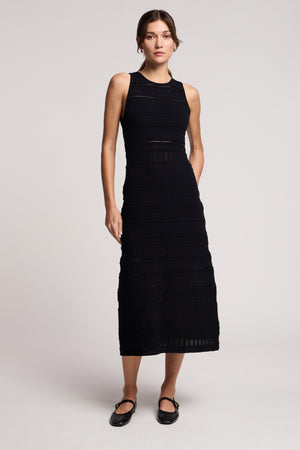 A woman stands against a plain background wearing the YLENA DRESS by Velvet by Graham & Spencer, an ankle-length, sleeveless, fitted A-line dress with subtle horizontal patterns, paired with black Mary Jane shoes.
