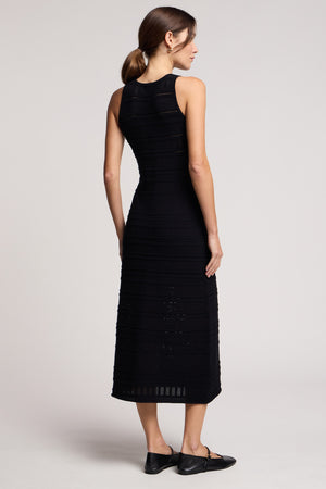 A woman with her hair in a low ponytail stands facing away, wearing the black YLENA DRESS by Velvet by Graham & Spencer—a sleeveless midi dress with an ankle-length hem—paired with black flats.