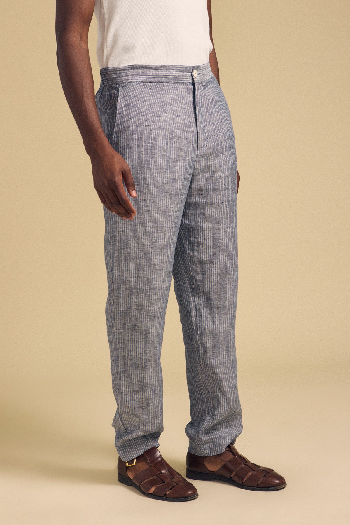 A person wears the JAE LINEN PANT by Velvet by Graham & Spencer—straight-leg striped linen pants with an elastic waistband—paired with a white sleeveless top and brown woven sandals, standing against a plain beige background.-41548475039937