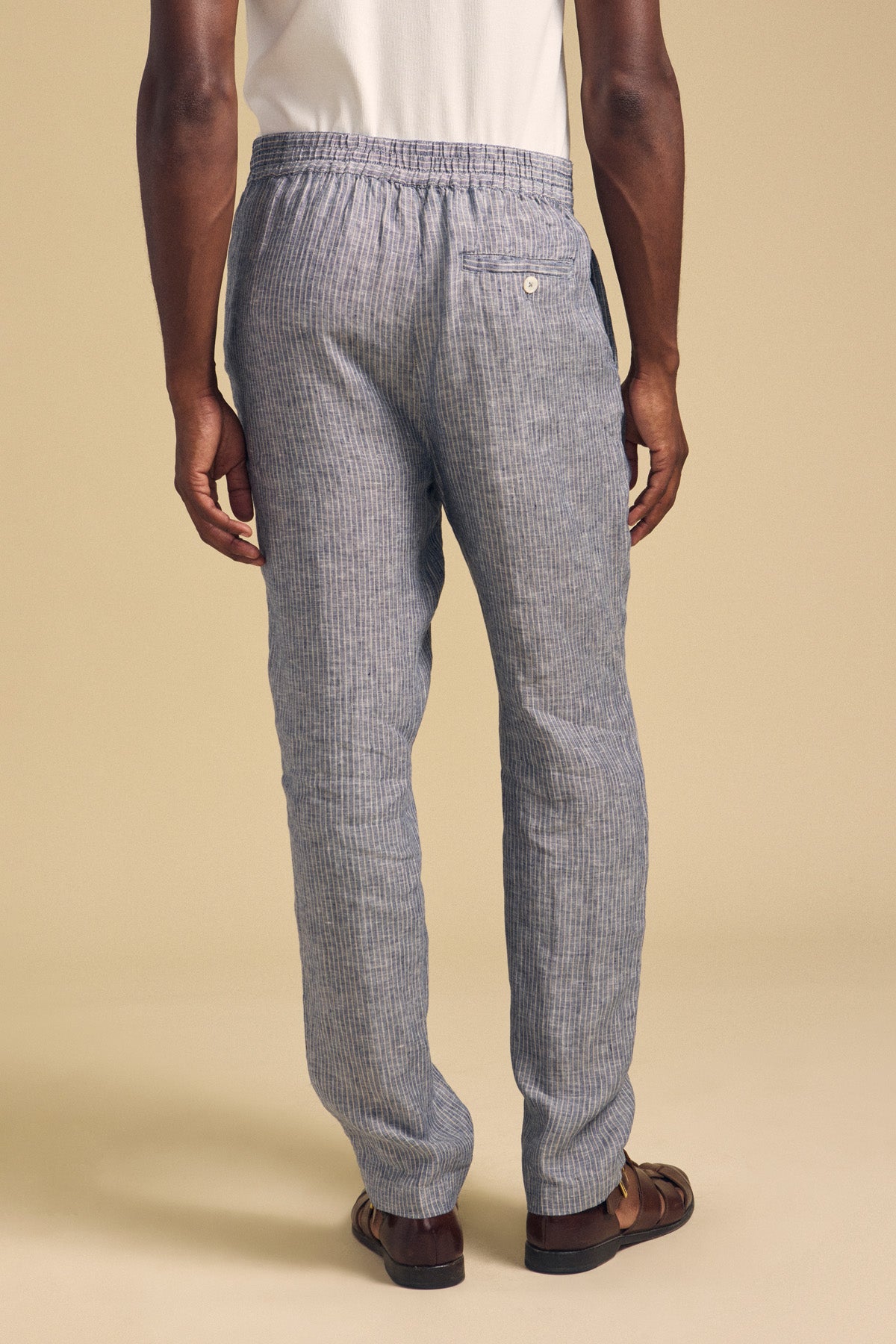 A person wearing Velvet by Graham & Spencer's JAE LINEN PANT in light blue and gray stripes, paired with a white shirt and brown sandals, stands facing away from the camera against a beige background.