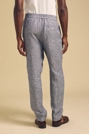 A person wearing Velvet by Graham & Spencer's JAE LINEN PANT in light blue and gray stripes, paired with a white shirt and brown sandals, stands facing away from the camera against a beige background.