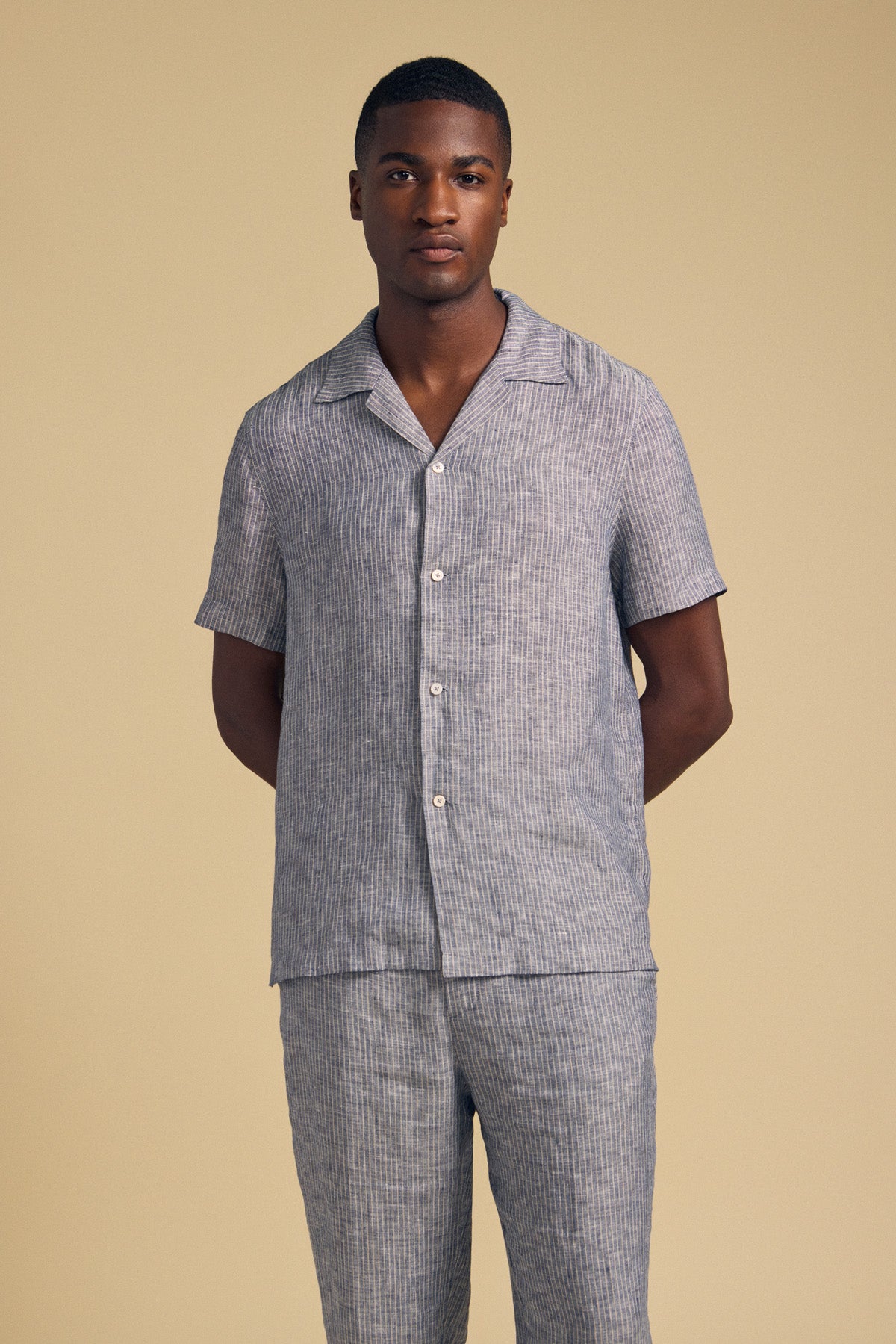 A man stands against a beige background wearing the JULIUS LINEN SHIRT by Velvet by Graham & Spencer, a short-sleeve striped linen button-up with a camp collar, paired with matching pants, his hands behind his back.-41548474384577